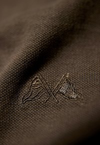 Mörkbrun tyg med en strukturerad väv och en broderad logotyp med korsade former, vilket framhäver en detaljrik brodyr på ytan.