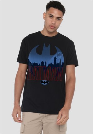 Young man wearing a black Batman t-shirt with city skyline and logo graphic, paired with beige cargo shorts, standing against plain background.