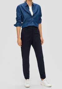 Tmavě modré kalhoty s přiléhavým střihem a délku do kotníků, v kombinaci s denimovou košilí na knoflíky. Bílá teniska dokončuje outfit.