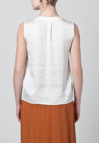 Hedvábný bezramínkový top v krémové barvě s jemným leskem, kulatým výstřihem a záhybem na zadní straně. Dcera s hnědou sukní.