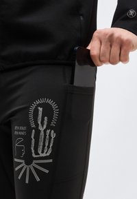 Main glissant un smartphone dans la poche noire d’un pantalon avec un graphisme de cactus et de visage et le texte « Open Roads Open Minds ».