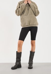 Fleece-hættetrøje i beige med en lomme, kombineret med sorte biker-shorts og knælange sorte støvler med metalaccenter og en kraftig sål.
