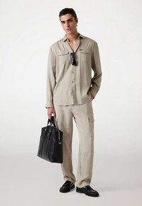 Camisa de lino beige con dos bolsillos en el pecho, combinada con pantalones cargo beige a juego. El modelo lleva un bolso tote de cuero negro y zapatos negros.