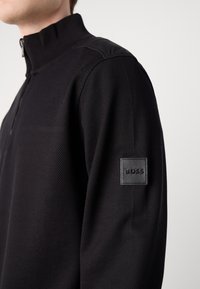 La felpa nera con zip presenta un corpo in maglia testurizzata, pannelli delle spalle lisci e una toppa in pelle con marchio in rilievo sul maniche.