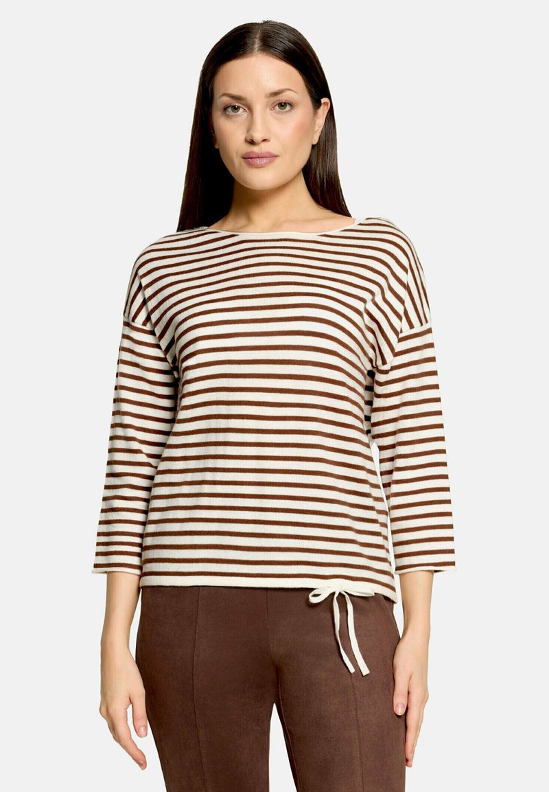 Femme portant un haut à manches longues marron et blanc à rayures horizontales avec un détail de noeud blanc, associé à un pantalon marron uni.