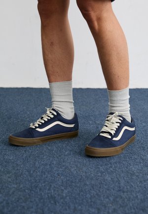 Zapatillas de ante azul marino con franjas laterales blancas, cordones crema y suelas de goma marrón, combinadas con calcetines grises de canalé sobre una alfombra azul.
