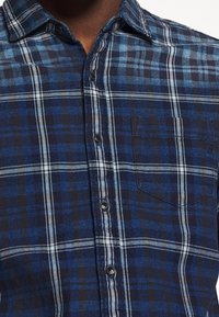 Primo piano di una persona che indossa una camicia a quadri blu e nera con taschino e colletto.