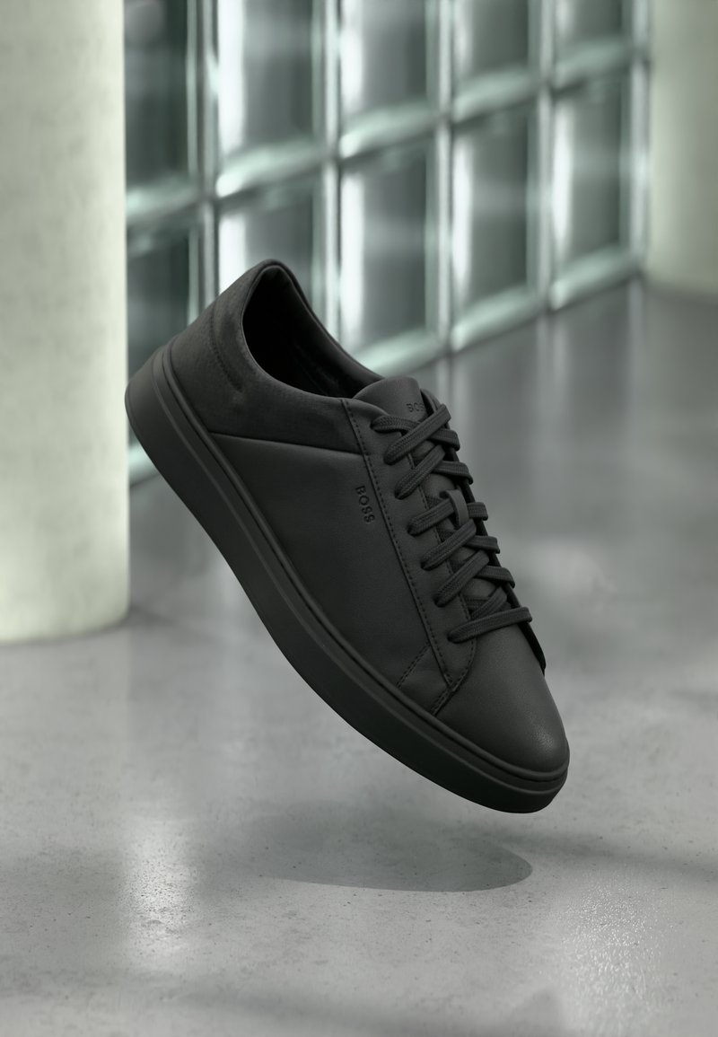 Baskets basses en cuir noir avec lacets et logo "BOSS" flottant au-dessus d'un sol en béton lisse près de blocs de verre.