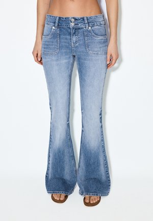 Svijetloplave traperice visokog struka širokih nogavica s prednjim džepovima, koje nosi osoba bez obuće koja stoji na bijeloj pozadini.