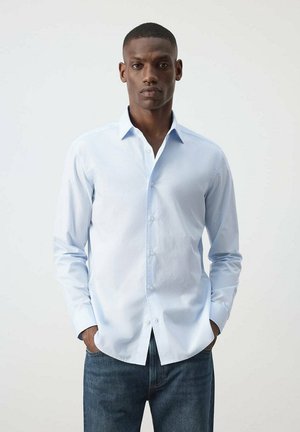 Hombre con una camisa azul clara abotonada y vaqueros azul oscuro, de pie contra un fondo blanco liso con las manos en los bolsillos.