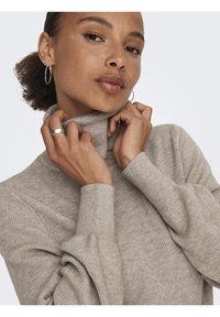 Beige polokrage-tröja med ribbad textur, långa ärmar och tighta manschetter. Modellen drar försiktigt i kragen och visar upp det mjuka tyget.