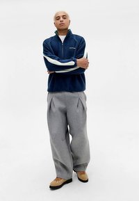 Tengerészkék polár cipzáras kabát fehér csíkokkal, szürke bő fazonú melegítőnadrággal és bézs fűzős cipőkkel, semleges háttér előtt állva.