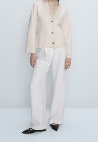Light pink knit cardigan with a deep V-neck, four front buttons, two pockets, worn over white wide-leg trousers and black pointed flats.