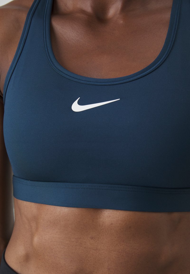 Top de sport court bleu foncé avec une large bande, présentant un logo Nike blanc à l'avant, fabriqué en tissu lisse et extensible.