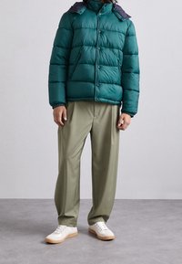 Grön puffjacka med marinblå krage, dragkedja och tryckknappar, i kombination med ljus olivfärgade byxor och vita sneakers. Slät, vadderad textur.