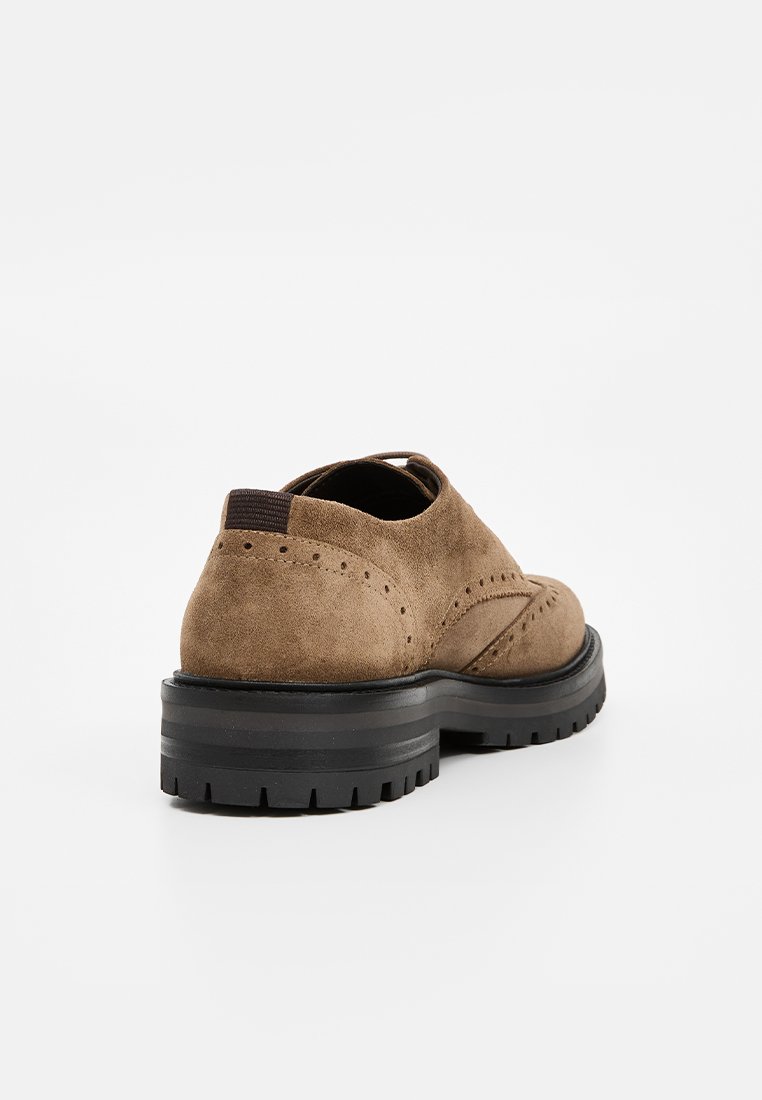 Brązowe zamszowe buty oxford z perforacjami, czarna gruba podeszwa z bieżnikiem oraz elastyczny uchwyt na pięcie, pokazane od tyłu na białym tle.