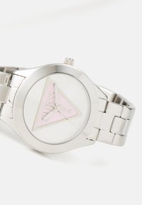 Montre en métal argenté avec un boîtier rond et un bracelet à maillons. Présente un cadran blanc avec un triangle rose et le logo de la marque. Aiguilles des heures et des minutes simples.