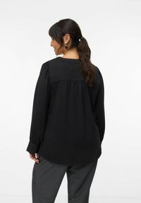 Blouse noire à manches longues avec un col rond, des épaules froncées et une coupe fluide, associée à un pantalon gris, présentant une texture lisse.