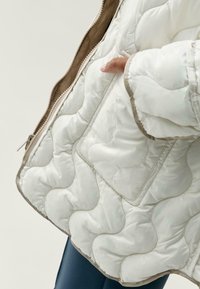 Bílá prošívaná bunda s vlnitým vzorem, béžovým lemováním a viditelným zipem. Obsahuje boční kapsu a hladkou, lesklou texturu.