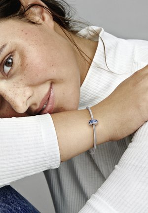 Bracciale d'argento con un accento di pietra blu, indossato su un polso, con uno sfondo di una maglia bianca a maniche lunghe texture.