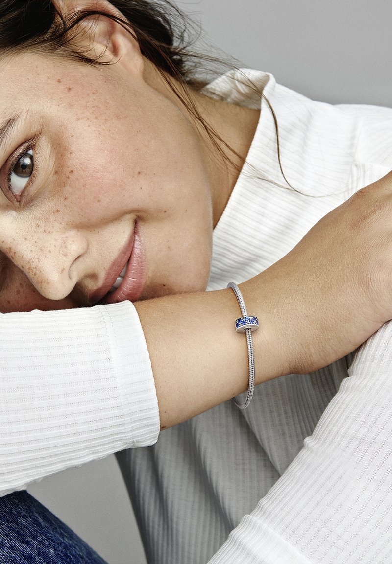 Pulsera de plata con un acento de gema azul, llevada en una muñeca, con un top blanco de manga larga texturizado en el fondo.