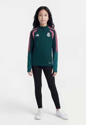 Fata stă așezată cu fața în față, purtând o jachetă sportivă Real Madrid verde închis și roz, colanți negri și adidași albi.