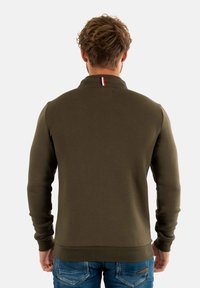 Olijfgroen sweatshirt met een hoge kraag, ribgebreide manchetten en een klein rood, wit en blauw accent bij de hals. Gladde textuur.