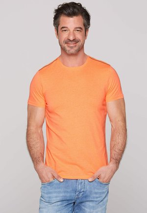 Oranje t-shirt met korte mouwen, gemaakt van zachte stof, met een ronde hals en zwarte accentstrepen op de schouders; gecombineerd met lichtblauwe jeans.