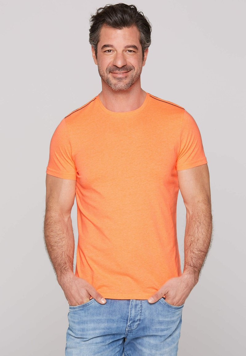 Oranje t-shirt met korte mouwen, gemaakt van zachte stof, met een ronde hals en zwarte accentstrepen op de schouders; gecombineerd met lichtblauwe jeans.