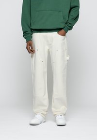 Pantalon cargo en coton blanc avec poches latérales et accents métalliques, associé à un sweat à capuche vert. Le look présente une coupe décontractée et des ourlets retroussés.