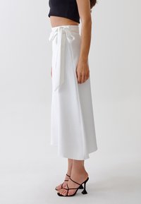 Jupe blanche avec un nœud à la taille, fabriquée dans un tissu doux. Conception évasée, longueur genou, associée à des talons noirs à lanières.