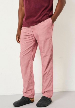 Mann mit rot-weiß gestreiften Hosen, burgunderfarbenem Hemd und dunkelgrauen Schlupfschuhen, der mit einer Hand in der Tasche steht.