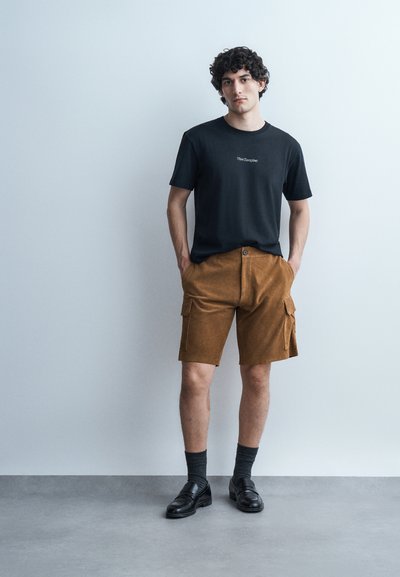 Jeune homme aux cheveux bouclés portant un t-shirt noir avec un logo, un short cargo marron, des chaussettes noires et des mocassins noirs, debout contre un mur uni.