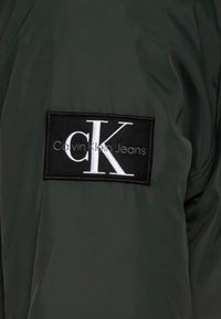 Calvin Klein grøn jakke med et fremtrædende sort stofmærke med "CK" og "Calvin Klein Jeans" i hvid skrift. Glat tekstur.