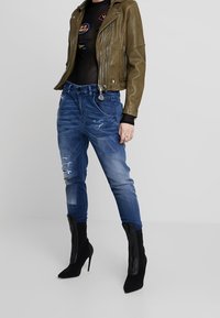 Olivově zelená kožená motorkářská bunda, černý síťový top s nášivkami, ošuntělé modré džíny, černé lodičky na jehlovém podpatku s texturovanými detaily.