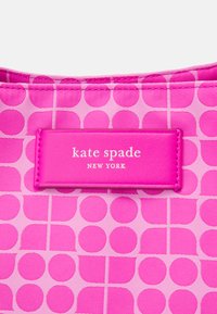 Un tessuto a fantasia rosa presenta cerchi sovrapposti. Un'etichetta rettangolare in pelle con la scritta "kate spade NEW YORK" è in evidenza.