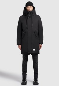 Jonge persoon in een zwarte winterparka met capuchon, zwarte beanie, slanke zwarte broek en zwarte laarzen, staande tegen een effen achtergrond.