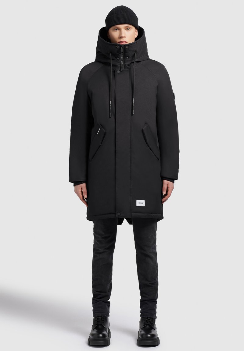 Jonge persoon in een zwarte winterparka met capuchon, zwarte beanie, slanke zwarte broek en zwarte laarzen, staande tegen een effen achtergrond.