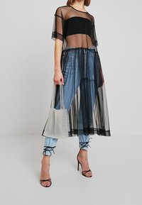 Žena v černých průhledných midi šatech přes černý bandeau top a modré džíny, držící bílou korálkovou kabelku a černé sandály na vysokém podpatku s šněrováním.