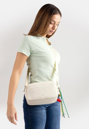 Donna che indossa una maglietta a maniche corte verde chiaro e jeans blu, con una borsa a tracolla bianca decorata con portachiavi colorati a nappina, che guarda verso il basso.