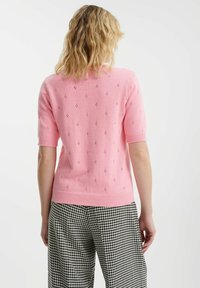 Jasnoróżowy sweter z krótkim rękawem z delikatnymi wzorami dziurek, z prążkowanym dekoltem i dołem, w połączeniu z spodniami w czarno-białą kratę houndstooth.