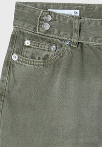 Gros plan sur un jean en denim vert montrant la ceinture, les boutons métalliques doubles, la poche avant avec rivets et l'étiquette de taille 36 à l’intérieur de la ceinture.