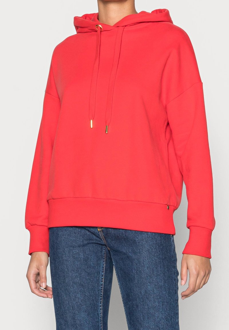 Rood hoodie gemaakt van zacht materiaal, met een koord met gouden metalen uiteinden, ribgebreide manchetten en een ontspannen pasvorm.
