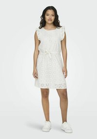 Weißes Lochmuster-Kleid mit Rüschensäumen, tailliertem Schnitt und ausgestelltem Rock mit floralem Ausschnittmuster. Getragen mit weißen Sneakers.