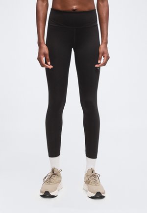 Persona con leggings ajustados negros, calcetines blancos y zapatos deportivos beige, de pie contra un fondo claro y liso.