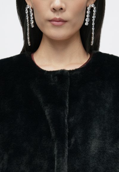 Crni kaput od umjetnog krzna s mekanom teksturom, bez ovratnika i s diskretnim tamnocrvenim unutarnjim oblogom. Dugi, sjajni kristalni naušnice.