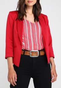 Blazer rouge ajusté à manches trois-quarts, associé à un haut rayé rouge et blanc, un pantalon noir, et une ceinture en cuir marron avec une boucle en argent.