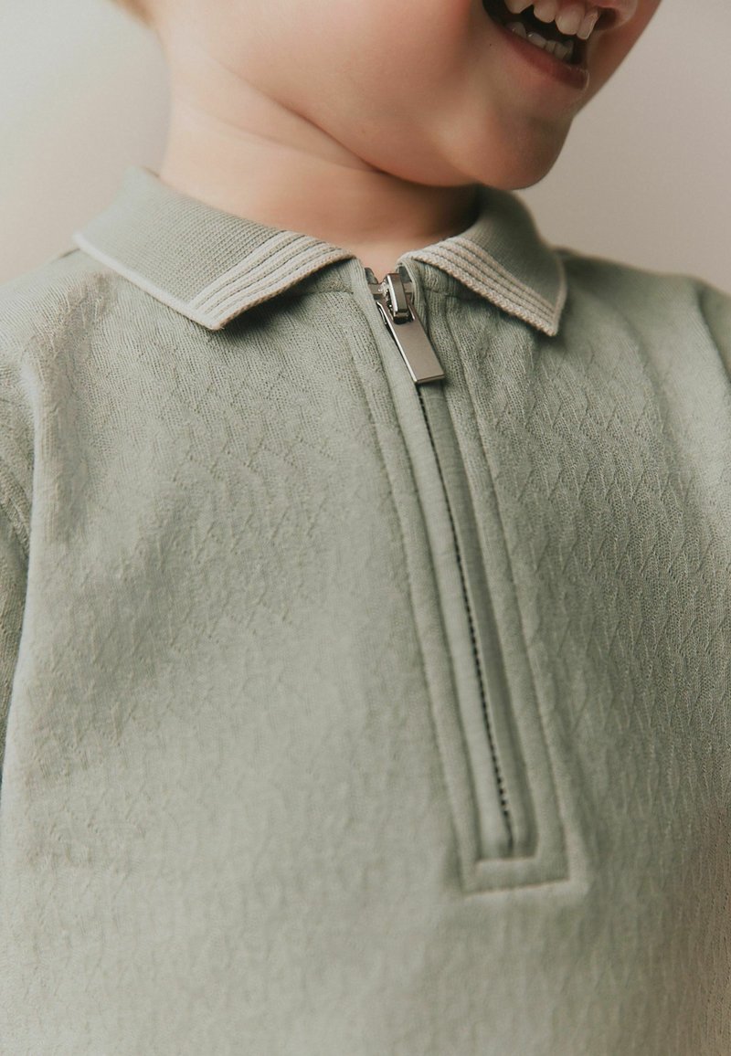 Niño con una camisa de cremallera texturizada color verde claro y cuello, sonriendo con la boca abierta y mostrando los dientes.