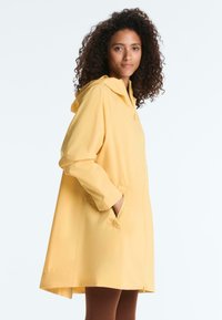 Femme aux cheveux bouclés portant un imperméable jaune ample avec une capuche, debout de côté avec une main dans la poche.