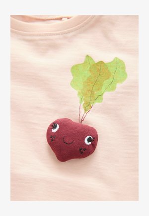 Svetloružová košeľa má plyšovú, maroon zeleninu s vyšitými očami a ústami. Zelený listový dizajn hore pridáva detail.